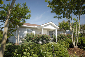 A small single-story white house is surrounded by green bushes and trees.