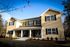 A white pathway leads to the wooden doors of a yellow two-story house with black shutters.