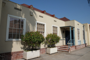 A concrete path leads to a one-story yellow building with a brick foundation and white latticed windows.