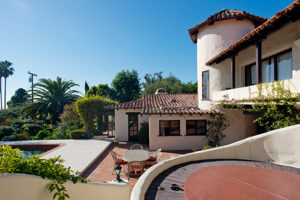 A Spanish-style home with three stories and a pool sits surrounded by palm trees, vines, and other greenery.