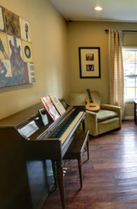 An old wooden piano sits on the left side of the room and a guitar rests on one of two tan chairs.
