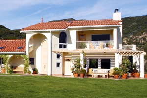 A white house with a red roof and an arched entryway sits on a large open field of grass.