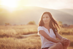 A woman sits in a field with her hands on her knee and the sun washing over her.